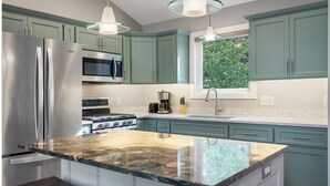 A modern kitchen with teal cabinets, stainless steel appliances, granite countertops, and pendant lighting.