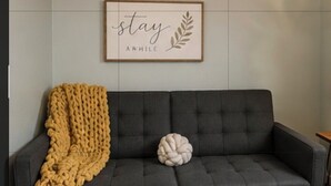 En mörkgrå soffa med en gul, stickad pläd och en vit, rund kudde, en inramad skylt med texten ”stanna kvar ett tag” och ett träpanelat tak.