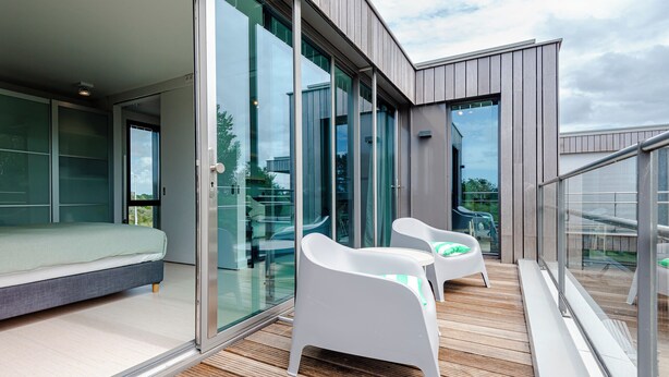 En moderne balkon med en seng, to hvide stole og en glasdør, der fører til et værelse.