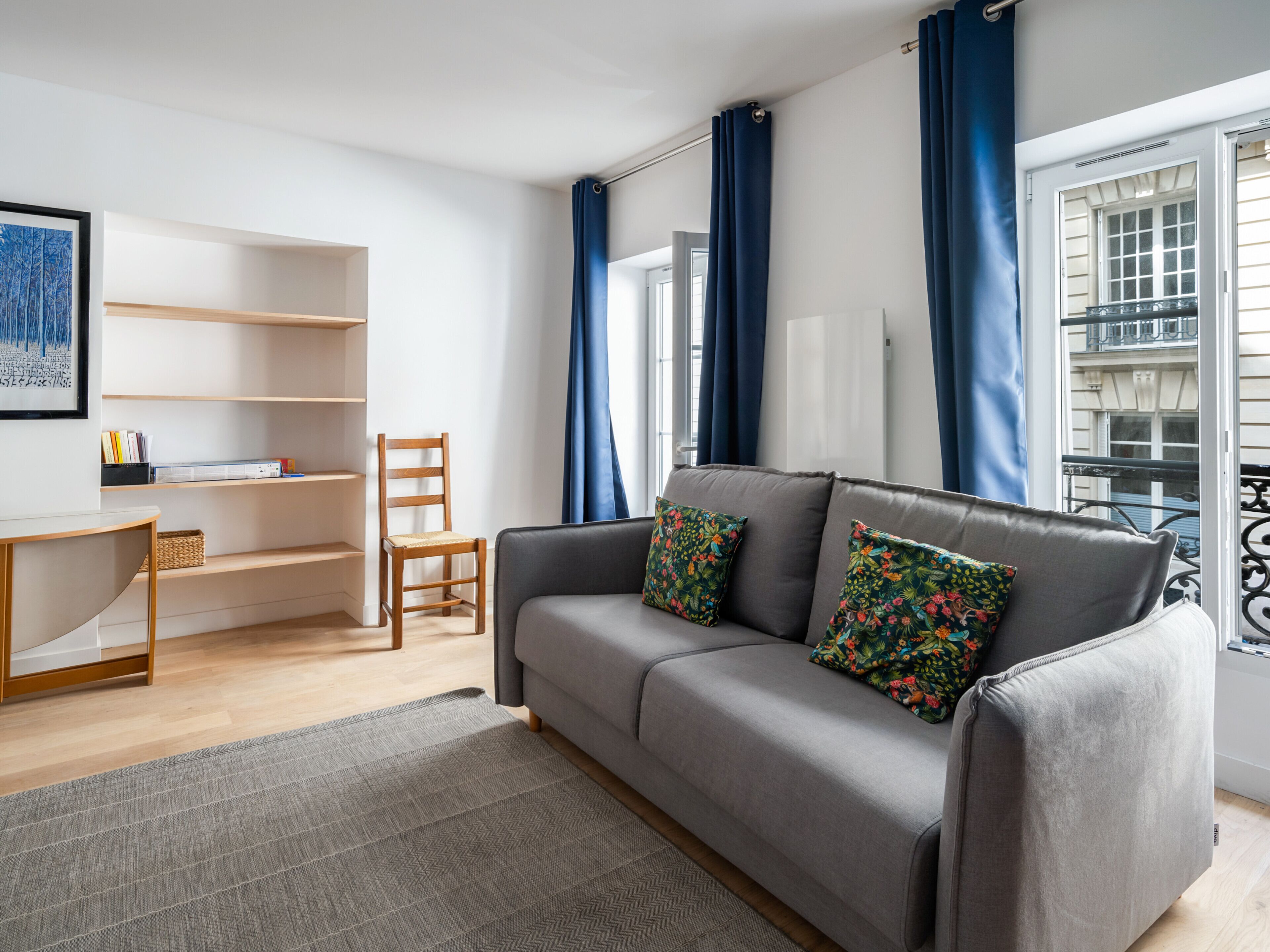 A living room with a grey sofa, two floral cushions, a wooden chair, a white desk with shelves, and a window with blue curtains.