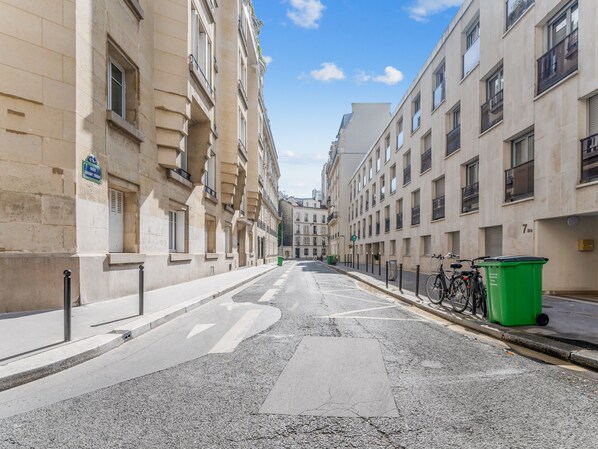 Une rue bordée de bâtiments des deux côtés, un vélo garé au bord du trottoir et une poubelle verte.