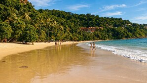 Una playa con un hotel al fondo, cielos despejados y aguas tranquilas.