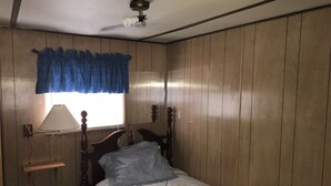 A bedroom with wooden paneling, a bed with a patterned bedspread, a nightstand with a lamp, and a window with a blue curtain.