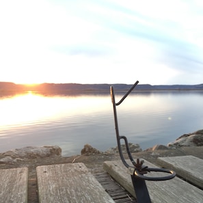 Eine hölzerne Bank mit einer Metallskulptur im Vordergrund, mit Blick auf einen stillen See bei Sonnenuntergang.