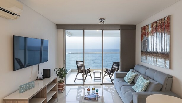 A modern living room with a large flat-screen TV, a grey sofa, and a balcony with a view of the sea. - Cartagena Home with a Heavenly View (Cartagena)
