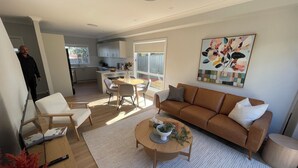 Een moderne woonkamer met een bruine bank, witte fauteuils en een salontafel. Er is een eethoek met een tafel en stoelen, en een keuken op de achtergrond.