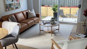 Een moderne woonkamer met een bruine leren bank, een ronde houten salontafel en een grijze fauteuil. Een groot raam biedt uitzicht op een houten terras en een bloempot.