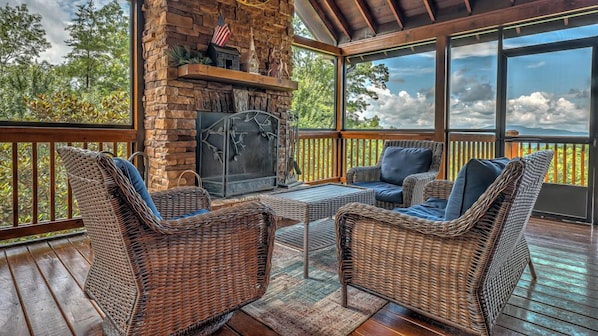 A covered outdoor area with wicker furniture, a stone fireplace, and large windows offering a view of trees and mountains.