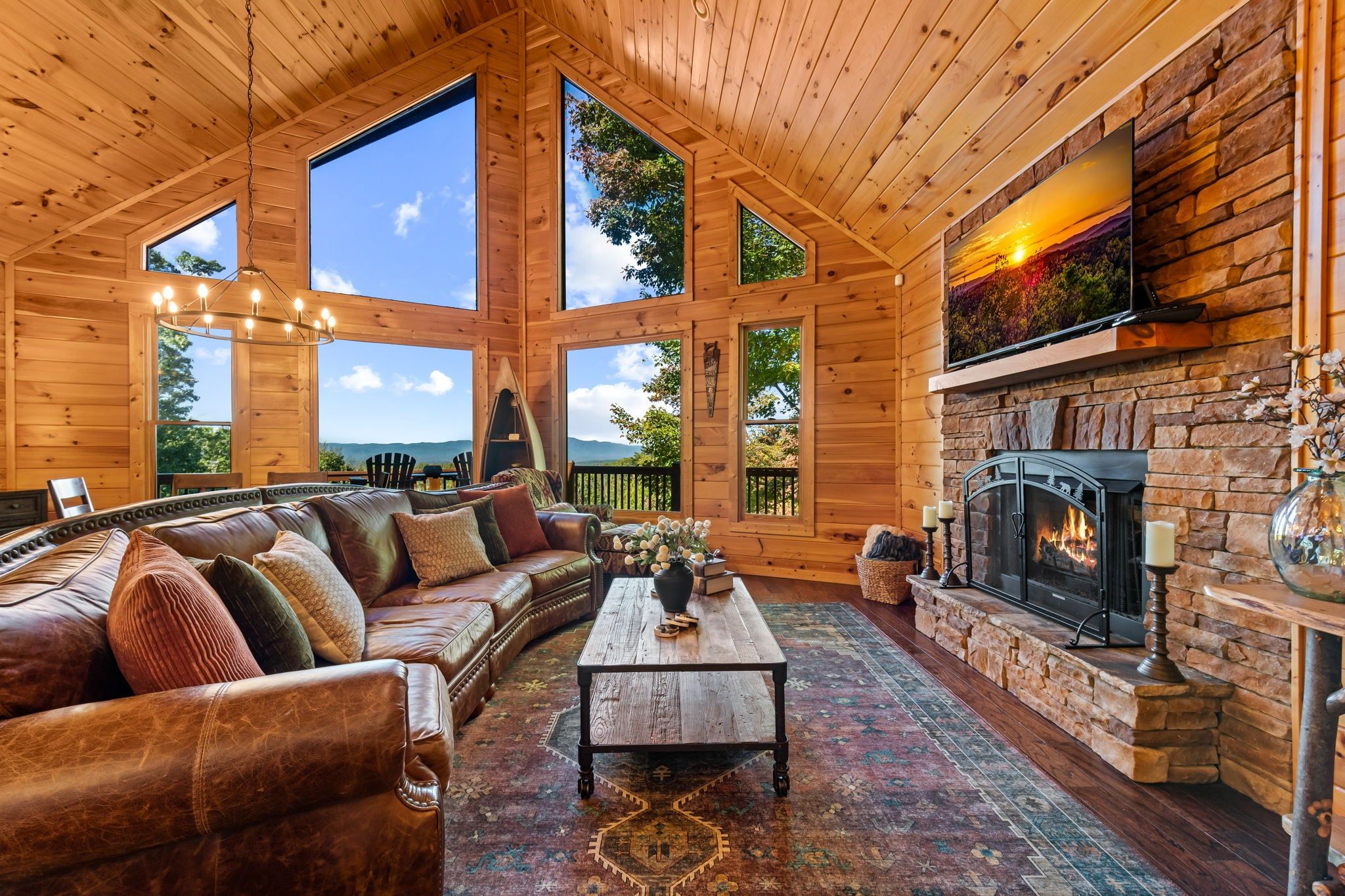 A log cabin interior with a large sofa, a fireplace, and large windows offering a view of the outdoors.