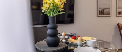 A modern dining area with a round glass table, a vase of yellow flowers, and a flat-screen TV on the wall.