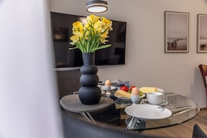 Un comedor moderno con una mesa redonda de vidrio, un florero con flores amarillas y un televisor de pantalla plana en la pared.