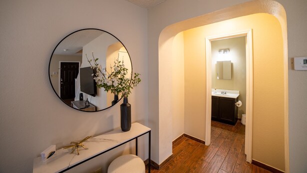 Un pasillo que conduce a un baño con un espejo redondo, un florero con flores y una consola con una lámpara.