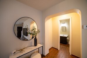 A hallway leading to a bathroom with a round mirror, a vase with flowers, and a console table with a lamp.