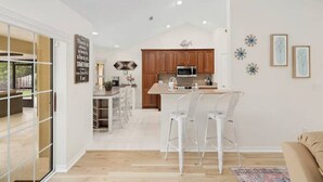 A modern kitchen with wooden cabinets, a central island with bar stools, and a dining area with a sofa. - Spacious Beachy Retreat with Heated Pool! Short walk to beach•Pet friendly•Bikes (St. Augustine)