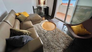 A modern living room with a leather sofa, a round wooden coffee table, and a dark armchair. A dining area with a table and chairs is visible through a glass door leading to a balcony with a view of the sea. - Holiday apartment with balcony and air conditioning (Crikvenica)