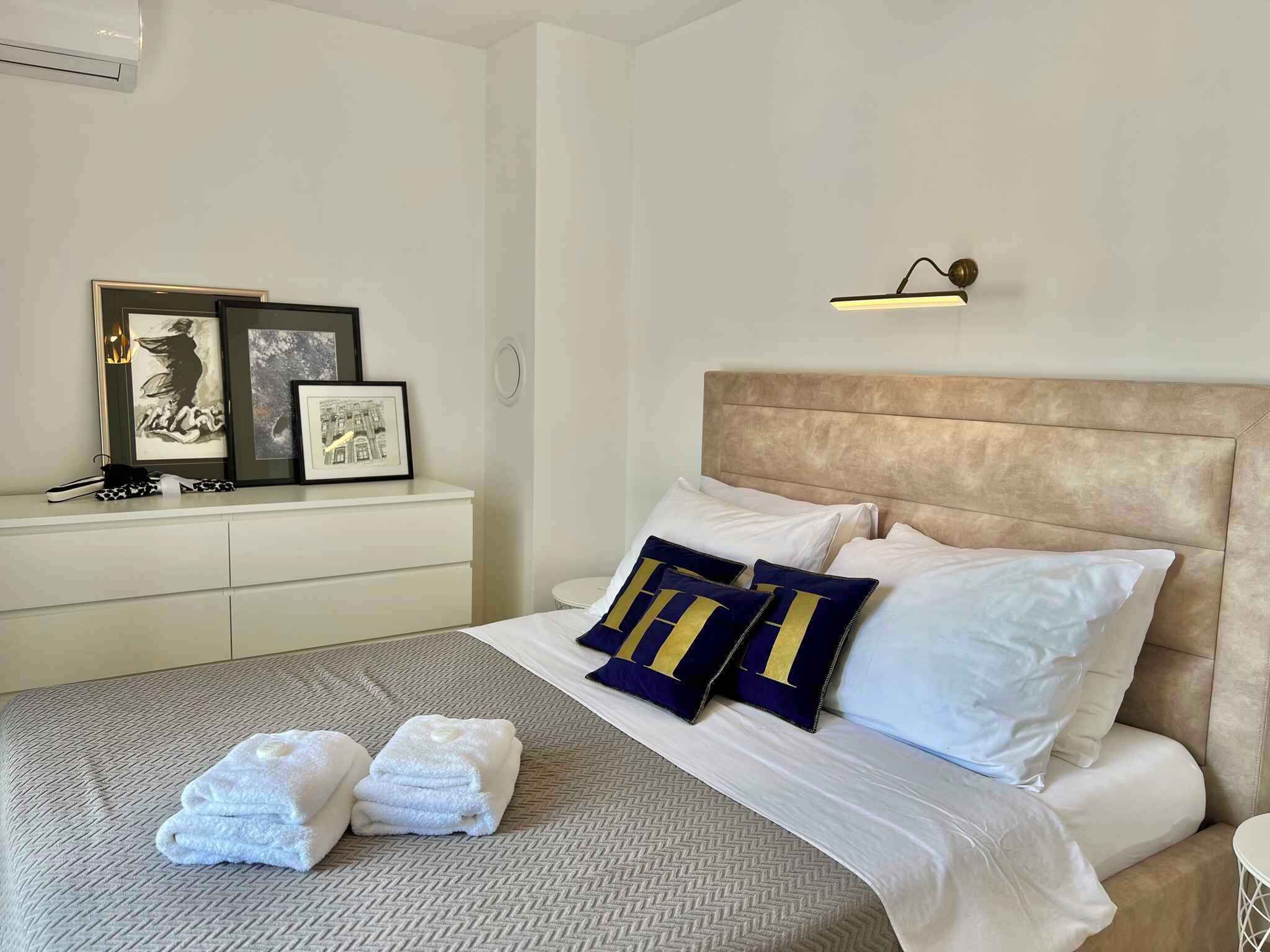 Una cama bien hecha con almohadas blancas y azules, un cabecero y una mesita de noche con cuadros enmarcados.