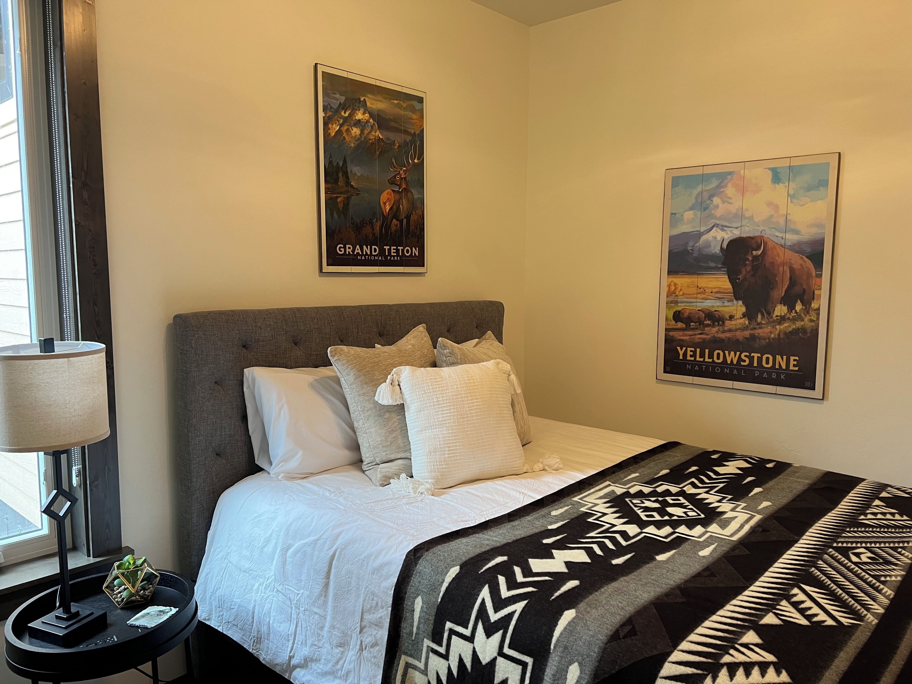 Une chambre à coucher avec un lit, des oreillers, une couverture, une table de chevet, une lampe et deux affiches représentant les parcs nationaux de Grand Teton et de Yellowstone.