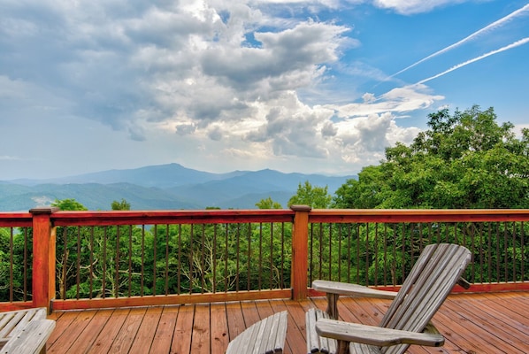 A wooden deck with a red railing, two wooden chairs, and a view of mountains and trees under a partly cloudy sky.