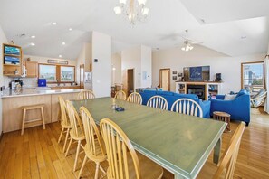 A spacious living area with a dining table and chairs, a blue sofa set, a television, and a kitchen area with wooden cabinets.