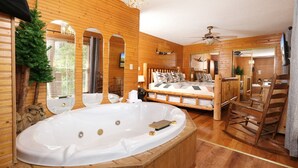 A wooden cabin interior with a large oval hot tub, a bed with multiple pillows, and a wooden rocking chair. - Cozy Bear Camp - Beautiful Views, Outdoor Hot tub, Firepit + More! (Sevierville)