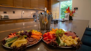 Une cuisine moderne avec des meubles en bois, un plan de travail sur lequel sont disposés deux plateaux garnis de fruits et de fromages variés, un réfrigérateur à vin et un vase rempli de fleurs.