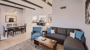 Un salon moderne avec un canapé gris foncé, un fauteuil bleu et une table basse en bois. On peut voir un ventilateur de plafond et des spots encastrés.