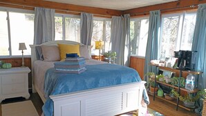 Ein Schlafzimmer mit Bett, Nachttischen, einer Lampe, einem Regal mit Pflanzen und einem Fenster mit Vorhängen.