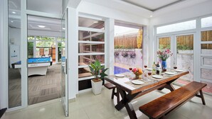 A dining area with a pool table, a long wooden table set for dining, and a view of a pool and garden through large windows. - Nelly Villa  | Copia Villa Hoi An (Thành phố Hội An)