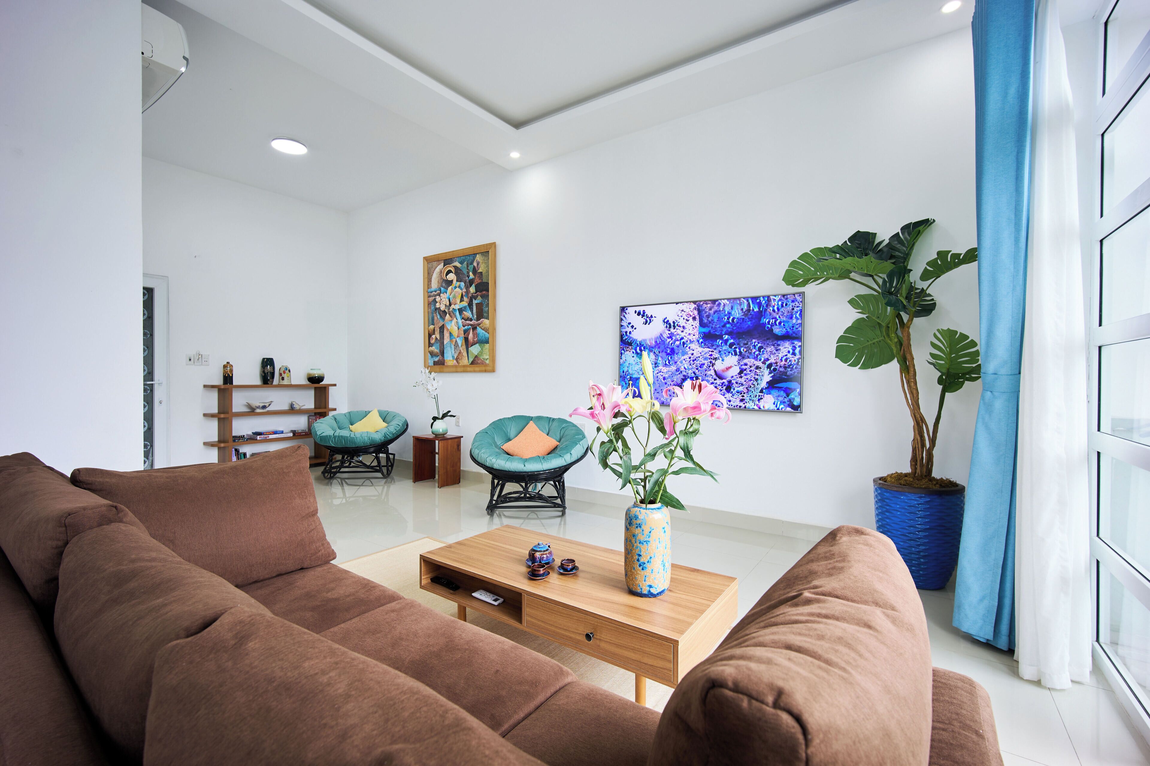 Un salon moderne avec un canapé marron, une table basse en bois, un vase bleu avec des fleurs, une grande plante en pot et des œuvres d’art accrochées aux murs.