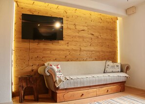 A cozy room with a wooden wall, a flat-screen TV mounted above a sofa, and a small wooden stool. - Charmante Ferienwohnung Handweberei mit Garten und Liegewiese, Wlan, 130 qm, Parkplatz (Saaldorf-Surheim)