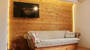 A cozy living room with a wooden wall, a flat-screen TV mounted above a sofa, and a small wooden stool. - Charmante Ferienwohnung Handweberei mit Garten und Liegewiese, Wlan, 130 qm, Parkplatz (Saaldorf-Surheim)