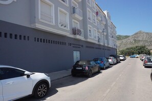 Une rangée de voitures garées le long d’une rue adjacente à un immeuble à plusieurs étages doté de balcons.