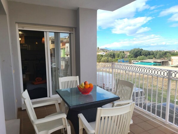 Un balcon avec une table en verre et des chaises blanches, un saladier rempli de fruits et une vue sur un quartier résidentiel.