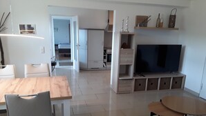 A modern living room with a wooden dining table, a sofa, a TV, and a kitchen area in the background. - Apartment "Bieniussa 3" with Private Garden & Wi-Fi (Gemünden am Main)