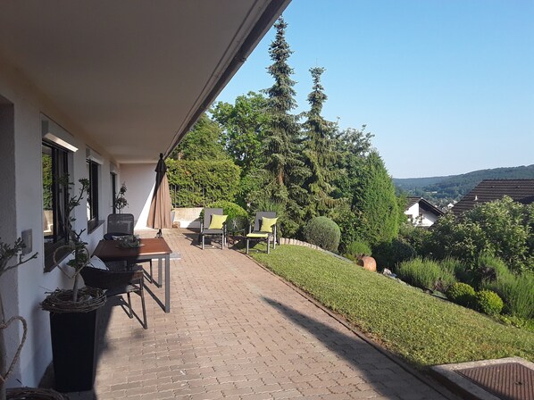 A covered outdoor patio with a table and chairs, potted plants, and a view of a residential area with trees and houses.