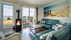 Una sala de estar con chimenea, un sofá verde, un sillón de mimbre y vistas a la playa a través de amplios ventanales.