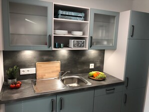 A kitchen with grey cabinets, a stainless steel sink, a wooden cutting board, and a fruit bowl.