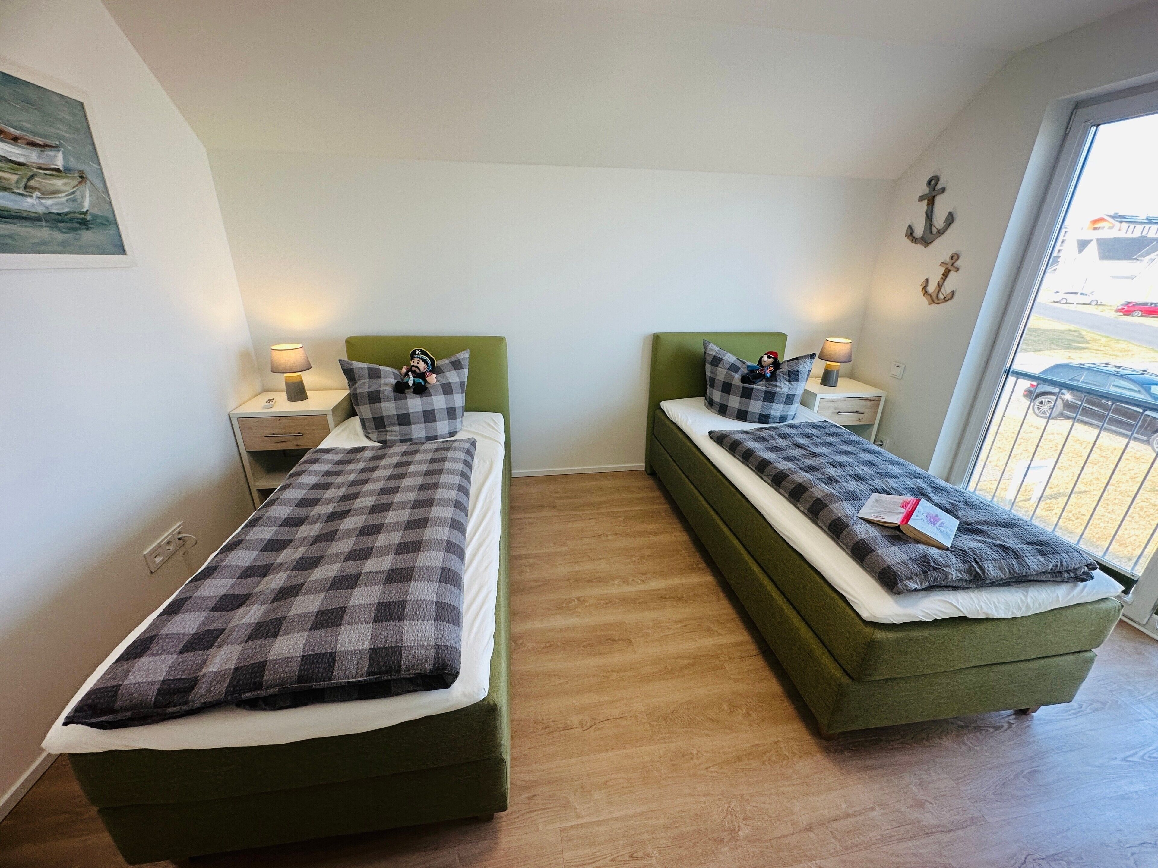 A twin room with two beds, each with a checkered blanket, green headboards, bedside tables with lamps, and a window with a view of parked cars.