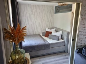 Ein kompaktes Bett mit Kissen, eine Vase mit Trockenpflanzen und eine hell gestrichene Wand mit dezenter Musterung.