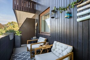 Eine Holzterrasse mit zwei gepolsterten Stühlen, einem kleinen Tisch und einem Fenster mit Blick auf Bäume.