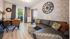 A modern living room with a brick wall, a large clock, a sofa, a dining table, and a view through a window. - Holiday Apartment 'Time 2 Sea' with Shared Garden, Balcony and Wi-Fi (Scharbeutz)