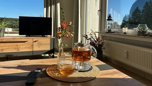 Uma mesa de madeira com um controle remoto, uma caneca de vidro com bebida, um vaso com flores e uma janela com vista para árvores e uma montanha.