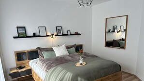 Quarto moderno com estrutura de cama de madeira, mesa de cabeceira, prateleira fixada na parede com fotografias emolduradas e um espelho.