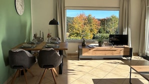Uma sala de jantar com uma mesa posta para quatro pessoas, um relógio na parede e uma televisão sobre um móvel.