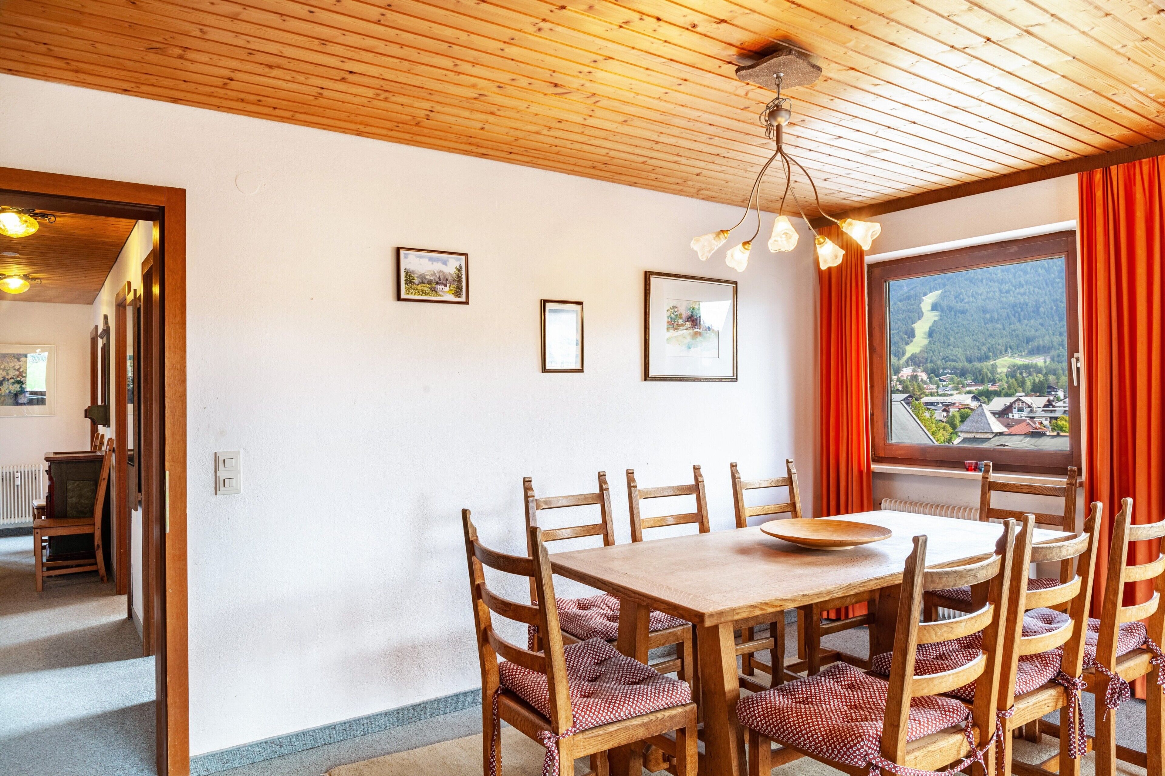 Un coin repas avec une table et des chaises en bois, des tableaux encadrés au mur et une fenêtre donnant sur une vallée.