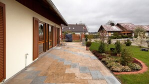 A covered patio with stone flooring, wooden shutters, and a garden with various plants and a small tree. - Apartment 'Haus Bross' with Balcony and Wi-Fi (Hayingen)