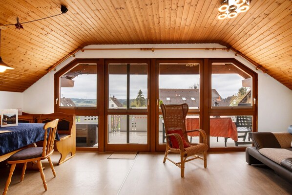 Une pièce spacieuse avec un plafond en bois incliné, de grandes fenêtres, une table à manger avec des chaises, un fauteuil en osier et un canapé.