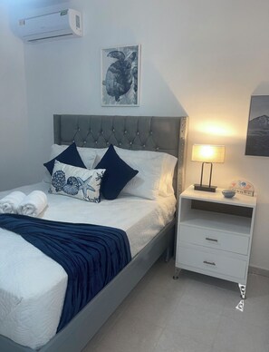 Una cama bien hecha con colcha y almohadas azul oscuro, una mesita de noche con lámpara, y un aire acondicionado fijado a la pared.