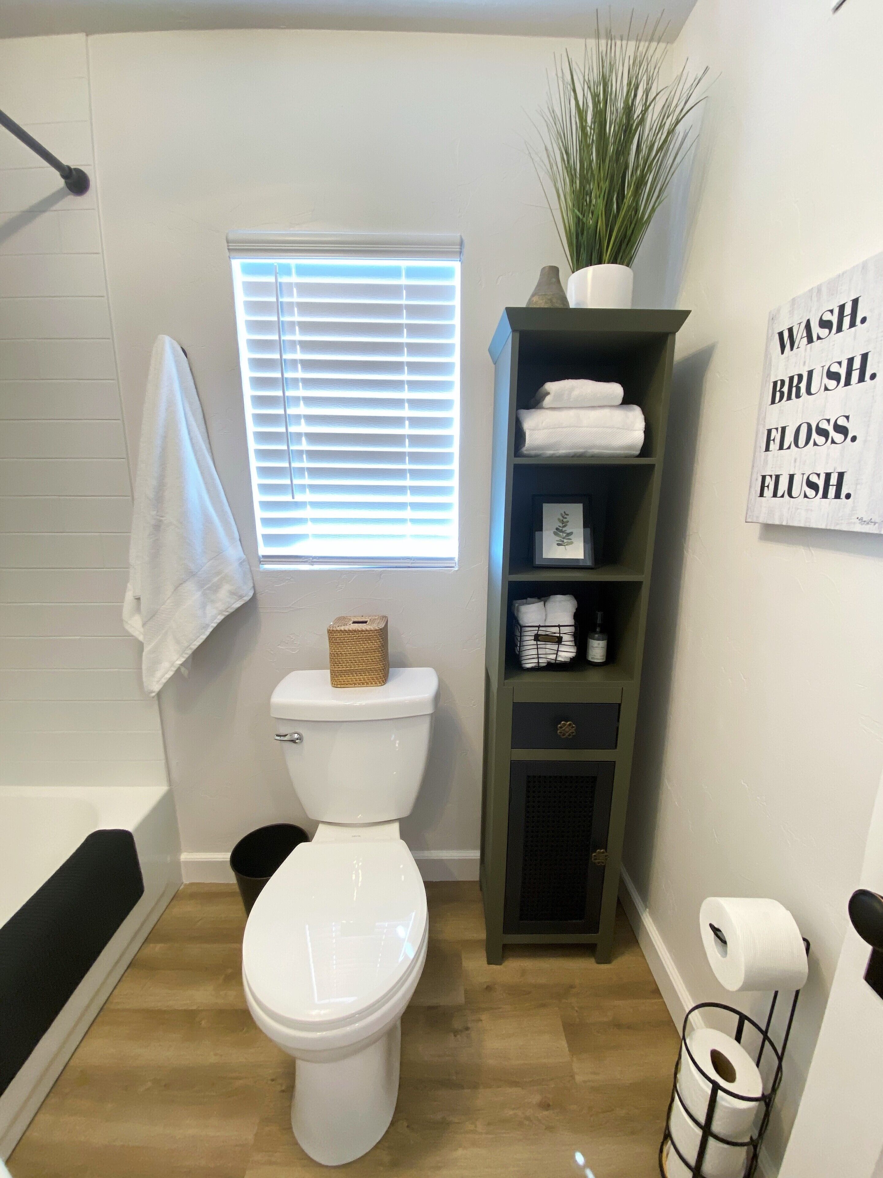 Ein Badezimmer mit einer weißen Toilette, einem Ablagefach für Toilettenartikel, einem Fenster mit Jalousien und einem Schild an der Wand.