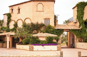 A building with arched windows, a tiled roof, and a garden with purple flowers.